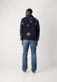 Σκούρο μπλε σακάκι με κεντημένα σχέδια, με υφή υφάσματος και μαλακό γιακά. Συνδυάζεται με ανοιχτό μπλε τζιν και λευκά παπούτσια.