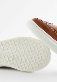 Chaussure en cuir marron avec un design perforé sur le dessus, dotée d'une semelle en caoutchouc blanche avec un motif de texture antidérapant.