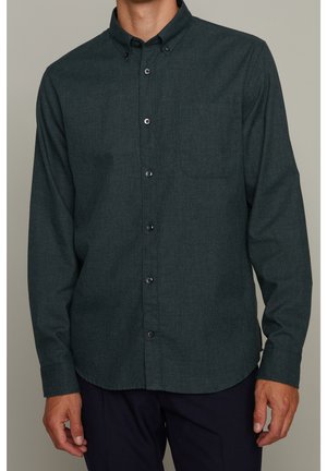 Homme portant une chemise boutonnée à manches longues vert foncé avec une poche poitrine et un col boutonné, associée à un pantalon foncé.