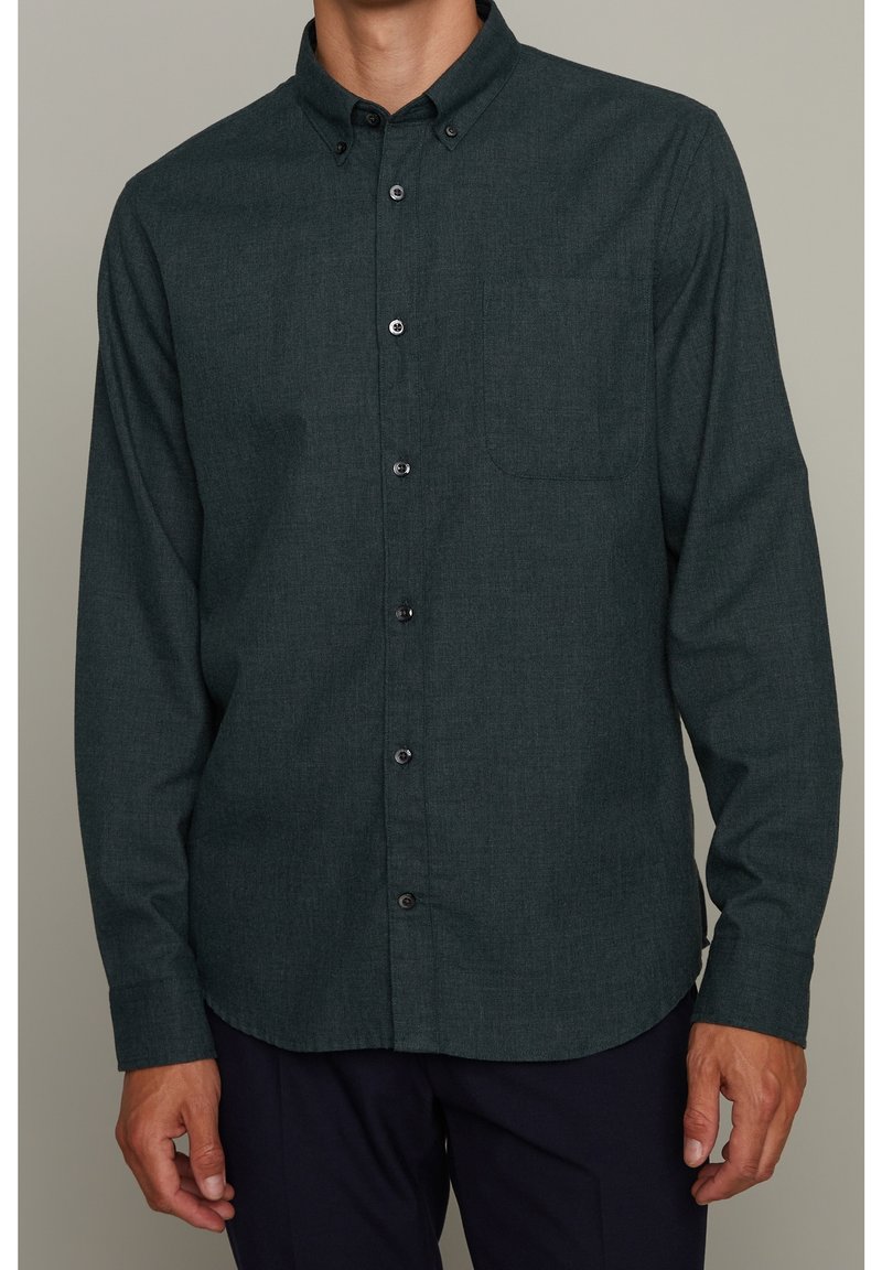 Homme portant une chemise boutonnée à manches longues vert foncé avec une poche poitrine et un col boutonné, associée à un pantalon foncé.