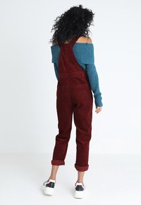 Kvinna med lockigt hår står vänd bort, klädd i en turkos off-shoulder tröja, vinröda uppvikta byxor och vita sneakers med svarta och röda detaljer.