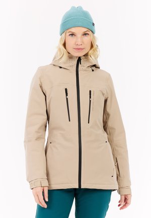 Beige wasserdichte Jacke mit Kapuze, mit Reißverschlusstaschen und schwarzen Akzenten. Strukturiertes Muster auf der Kapuze, getragen über angrünlichen Hosen.