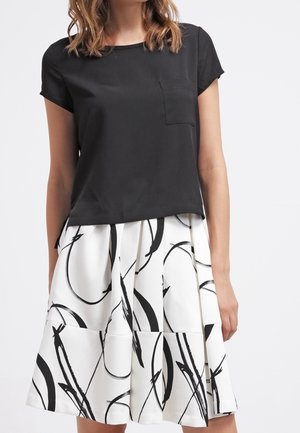Femme portant un haut noir à manches courtes avec une poche, associé à une jupe blanche présentant de grands motifs abstraits en coups de pinceau noirs.