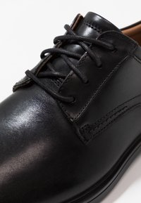 Chaussure en cuir noir avec une finition lisse, dotée de lacets ronds, d'accents cousus et d'un design classique. Vue partielle de la tige.