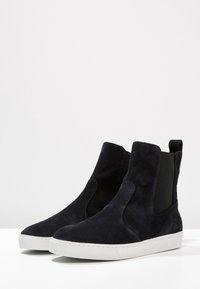 Bottines noires en daim avec des panneaux élastiques sur les côtés, semelle en caoutchouc blanche et texture lisse. Présente un design minimaliste et une silhouette épurée.