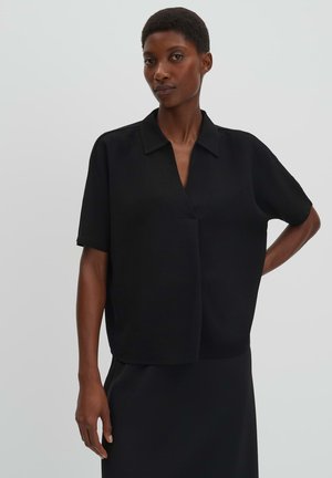 Zwart kort mouwen blouse met kraag, gecombineerd met een zwarte rok, gedragen door een staande persoon tegen een effen lichtgrijze achtergrond.