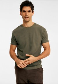 Khaki grøn kortærmet t-shirt med et struktureret, gitterlignende mønster og en rund halsudskæring; modellen står med hænderne foldet om livet.