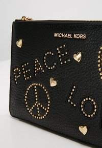 Pochette en cuir noir texturé avec des clous dorés, des accents en forme de cœur et un texte en relief "PAIX" et "AMOUR". Fermeture éclair sur le dessus.