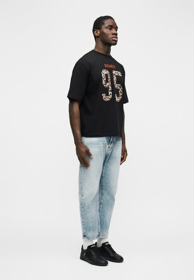 Άν δρας στέκεται στραμμένος προς τα δεξιά φορώντας μαύρο μπλουζάκι με λεοπάρ τύπωμα "95" και ανοιχτό μπλε τζιν με μαύρα αθλητικά παπούτσια και λευκές κάλτσες.