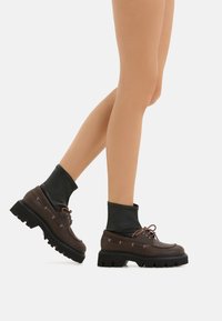 Scarpe in pelle marrone con una sezione per la caviglia in gomma nera, suola spessa e chiusura con lacci, dotate di occhielli in argento e una texture liscia.