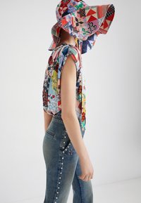 Giovane che indossa una canottiera a patchwork colorata e un cappello a falda larga coordinato, abbinato a jeans blu con borchie, in posizione di profilo.