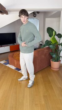 Vihreä neule-cardigan, vetoketjullinen malli, yhdistetty valkoisiin housuihin ja hopeisiin urheilukenkiin. Taustalla on kasvi ja oranssi sohva.