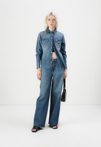Denim shirt met twee borstzakken en contrasterende stiksel, gecombineerd met wijde spijkerbroeken in een middelmatige wassingen, aangevuld met zwarte haksandalen.