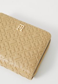 Portefeuille beige texturé avec un motif géométrique en relief, présentant un accent logo doré et une fermeture éclair sur le bord.
