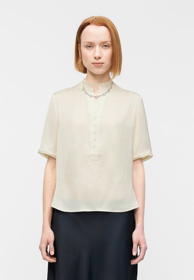 Blouse crème à manches courtes, patte de boutonnage et encolure décorative. Tissu léger et transparent avec une texture lisse.