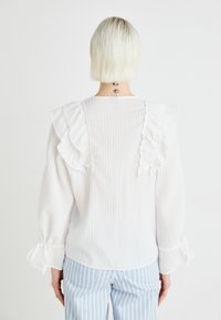 Hvit volangbluse med lange ermer, vertikal stripe tekstur og knyttede mansjetter, båret med blå og hvite stripete bukser; bakfra.