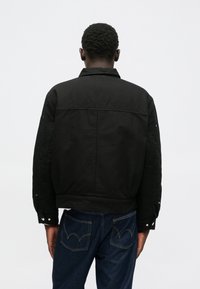 Chaqueta negra con cuello, hecha de una tela texturada, que presenta un corte relajado y puños con botones a presión, combinada con jeans azul oscuro.