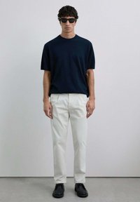 Suéter de punto azul marino, pantalones rectos blancos y zapatos negros. El modelo lleva gafas de sol oscuras. Diseño simple, textura suave, sin patrones.