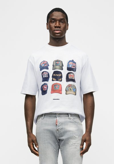 Άνδρας που φοράει λευκό μπλουζάκι με εννέα πολύχρωμα σχέδια από καπέλα και ανοιχτό γκρι τζιν με ραβδώσεις, στέκεται μπροστά σε απλό φόντο.