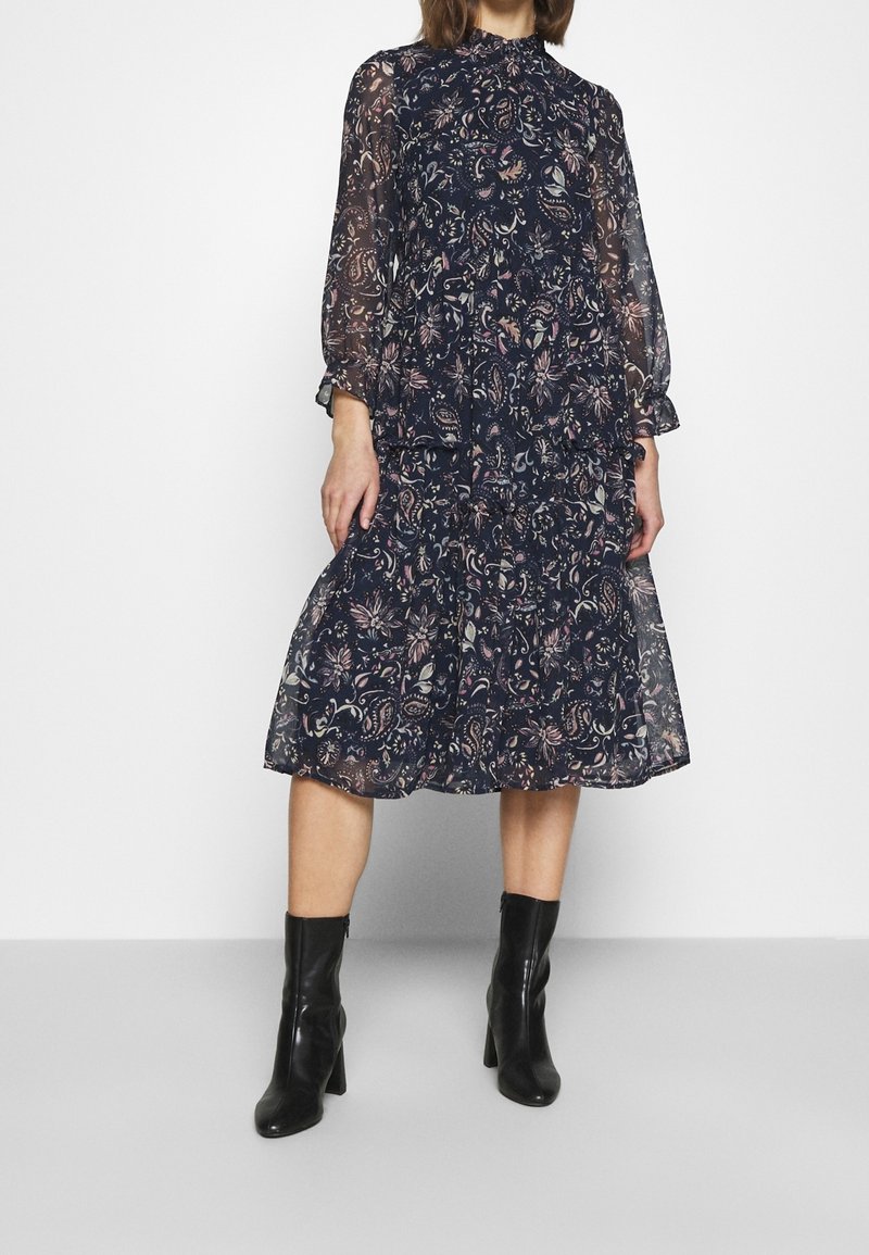 Femme portant une robe fleurie sombre longueur genou avec des manches longues transparentes, associée à des bottines noires à talons.
