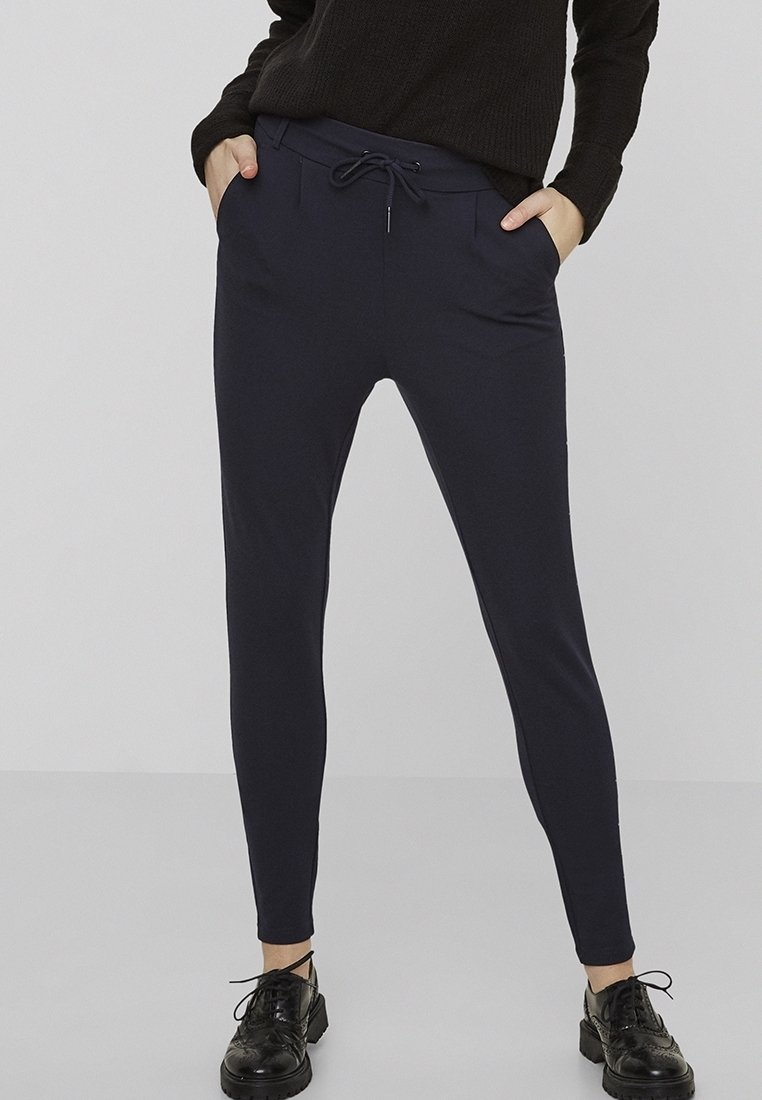Personne portant un pantalon noir slim à cordon de serrage, un pull noir et des chaussures noires à lacets, debout avec les mains dans les poches.