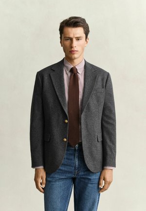 Mann mit dunklem Haar, der einen grauen Blazer, eine braune Krawatte, ein gestreiftes Hemd und blaue Jeans trägt, steht vor einem schlichten hellen Hintergrund.