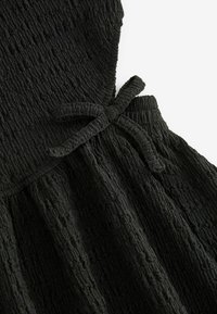 Détail d’une robe en tissu texturé noir avec taille froncée et nœud en tissu au centre devant.