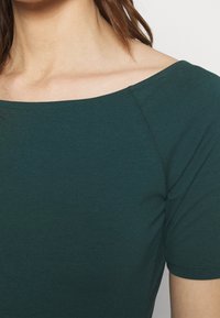 Mørkegrøn, tætsiddende top med en bred, rund udskæring og korte ærmer. Glat tekstur og diskret syning omkring kanterne.