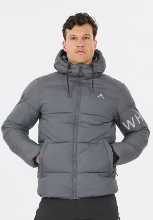 Mann trägt eine graue Steppjacke mit Kapuze, Hände in den Taschen, steht vor einem schlichten hellen Hintergrund, weißer ""WH""-Text am Ärmel.