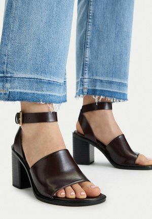 Füße in dunkelbraunen Leder-Sandalen mit Blockabsatz und Knöchelriemen, kombiniert mit hellblauen Jeans mit ausgefranstem Saum.