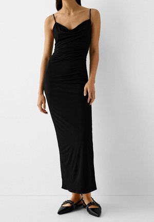 Femme portant une longue robe noire sans manches avec de fines bretelles et des ballerines noires pointues avec des brides cloutées, debout devant un fond blanc uni.