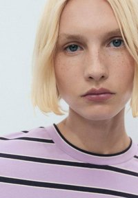 Modelka se světle blond vlasy a pihami, oblečená ve fialově pruhované košili s kulatým výstřihem. Pozadí je neutrální a minimalistické.