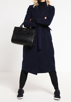 Cappotto navy con cintura, abbinato a pantaloni neri e borsa nera testurizzata. Presenta sneakers con cinturini e una silhouette elegante.