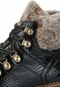Botas de charol negro con un collar de pelo sintético, ojales dorados y cordones negros, que presentan una superficie texturizada y una suela marrón claro.