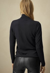 Schwarzes Langarmoberteil mit hohem Kragen und geripptem Muster, von hinten gezeigt. Kombiniert mit figurbetonten schwarzen Lederhosen.