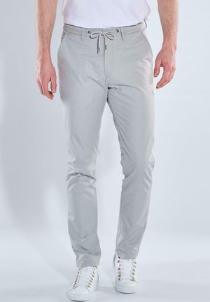 Mann trägt hellgraue Kordelhose und weiße Turnschuhe, steht vor einfarbigem Hintergrund.