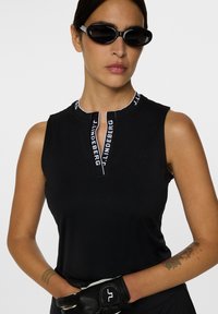 Svart utan ärmar topp med krage och dragkedja, med vita märkeskännetecken längs kragen. Strukturerad tyg och kroppsnära design.