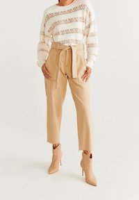 Frau in creme- und beige gestreiftem Strickpullover, hochtaillierten beigen Hosen mit Gürtel und beigen spitzen Absatzstiefeletten.