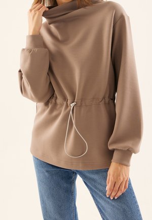 Mujer ajustándose el cuello de una camiseta de manga larga color topo con cintura ajustable mediante cordón, combinada con vaqueros azules.