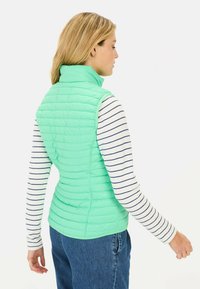Mintgrüne gesteppte Weste mit hohem Kragen, leichtem Design, Seitennähten und taillierter Form, getragen über einem dunkelblau-weiß gestreiften Langarmshirt.