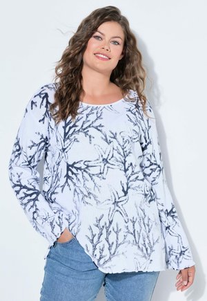 Kobieta o kręconych włosach, ubrana w biały sweter z czarnym wzorem korala i rozgwiazdy oraz niebieskie dżinsy, uśmiecha się z ręką w kieszeni.