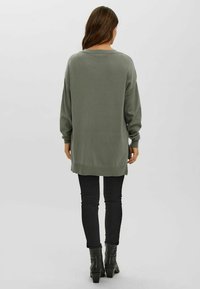 Olivgrüner, übergroßer Pullover mit langen Ärmeln, geripptem Saum und seitlichen Schlitzen, kombiniert mit schwarzen Skinny-Jeans und Stiefeletten.