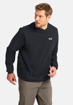 Uomo che indossa un maglione Jack Wolfskin nero a maniche lunghe e pantaloni marrone chiaro, guarda verso sinistra con un'espressione concentrata.