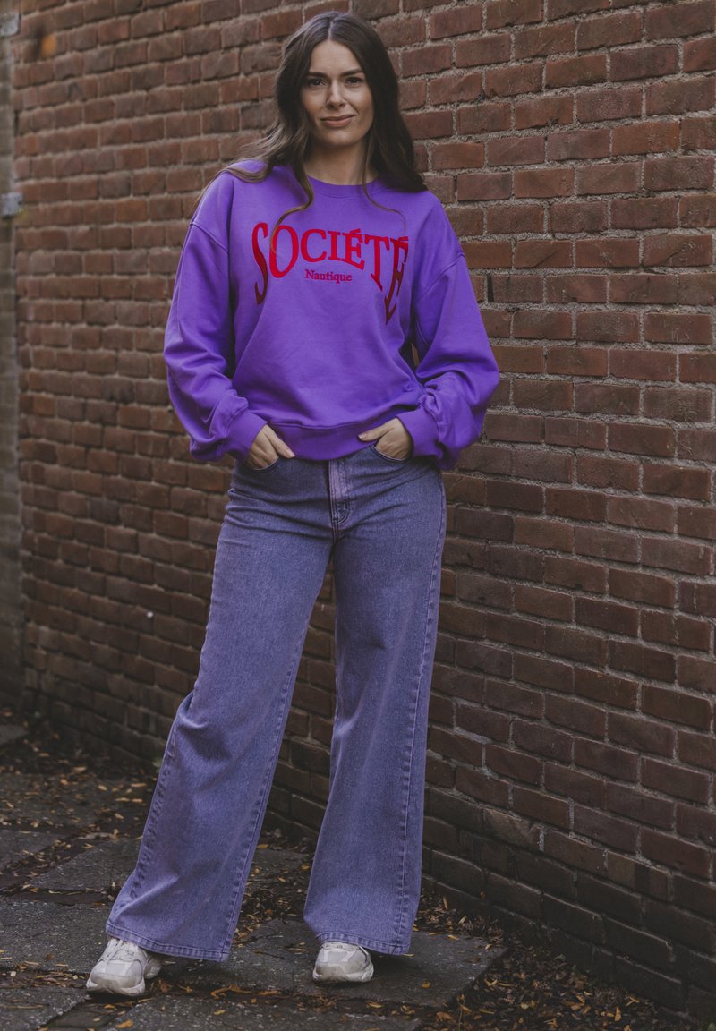 Paarse sweatshirt met rode tekst "Société Nautique," gecombineerd met lichtblauwe wijde jeans en grijze sneakers, voor een bakstenen muur.