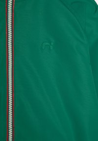 Grön zip-jacka med texturerad tyg, kontrasterande vit och röd randig dragkedja samt ett präglat logotyp på vänster sida.