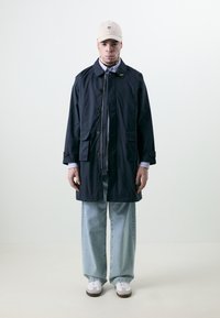Navy blue trench coat with button closure, two front pockets, and adjustable sleeves. Worn over a light blue shirt and relaxed-fit jeans.