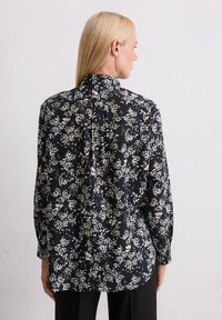 Femme aux cheveux blonds raides portant un blouse noire ample avec des motifs floraux blancs et jaunes, de dos contre un mur blanc uni.