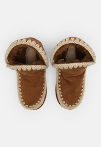 Pantofole in suede marrone con rivestimento in pelliccia, caratterizzate da un bordo intrecciato di colore crema attorno all'apertura e ai bordi. Tessuto morbido con una forma arrotondata.