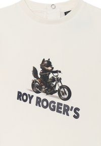 T-shirt color crema con un lupo cartoon in casco che guida una moto, con testo in grassetto che dice "ROY ROGER'S" sotto il grafico.
