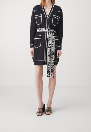Un long cardigan noir avec des contours de poches blanches et une longue ceinture portant l'étiquette "KARL LAGERFELD", porté avec des talons noirs à lanières.